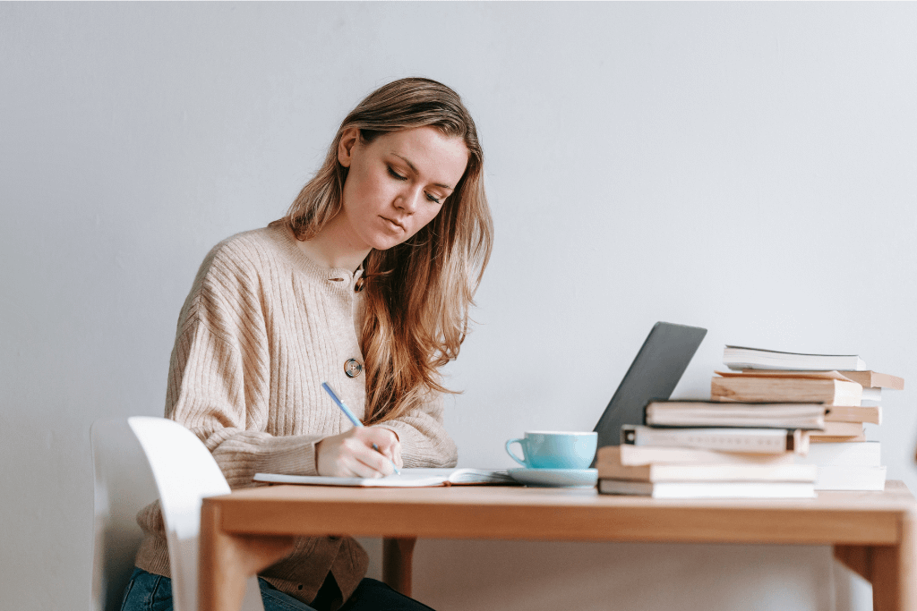 Concentrating woman writing ideas in notebook near laptop and books