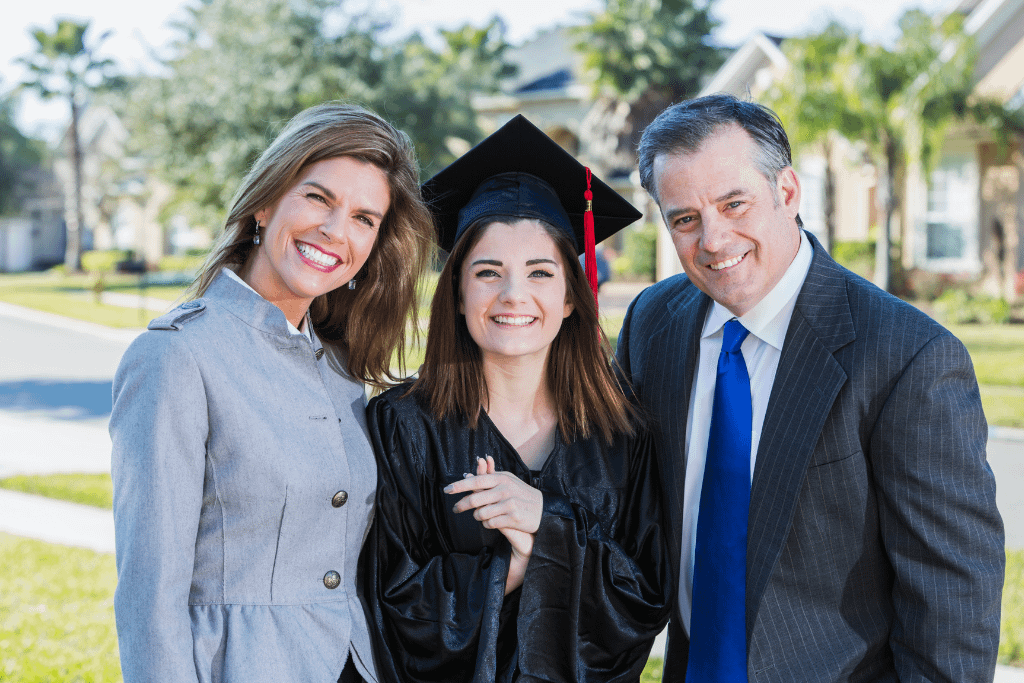 A teenage girl graduating from high school, posing with her parents, wearing a black cap and gown, smiling at the camera