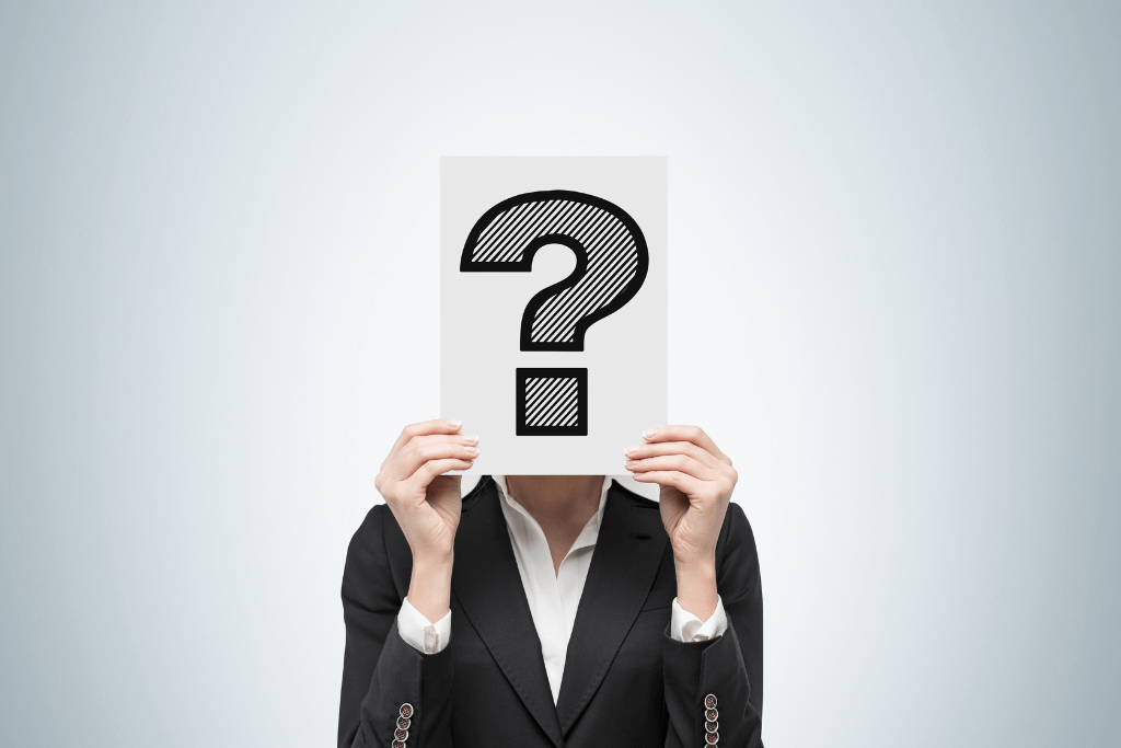 A person in a business suit holding a white sign with a large black question mark in front of their face, symbolising uncertainty or decision-making.