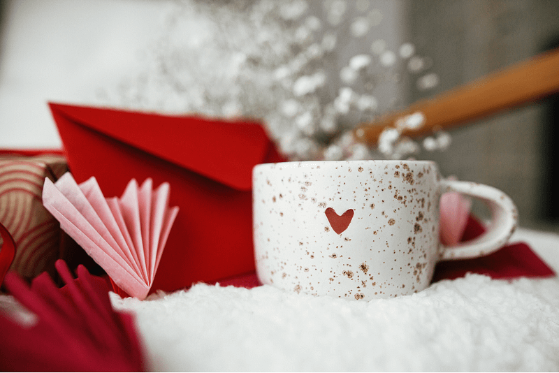 A cozy Valentine's Day-themed scene featuring a speckled ceramic mug with a small red heart design on its side. The mug is placed on a soft white surface, surrounded by romantic elements, including a red envelope, delicate pink paper origami, and baby's breath flowers in the background. The warm and inviting atmosphere suggests self-care and relaxation.